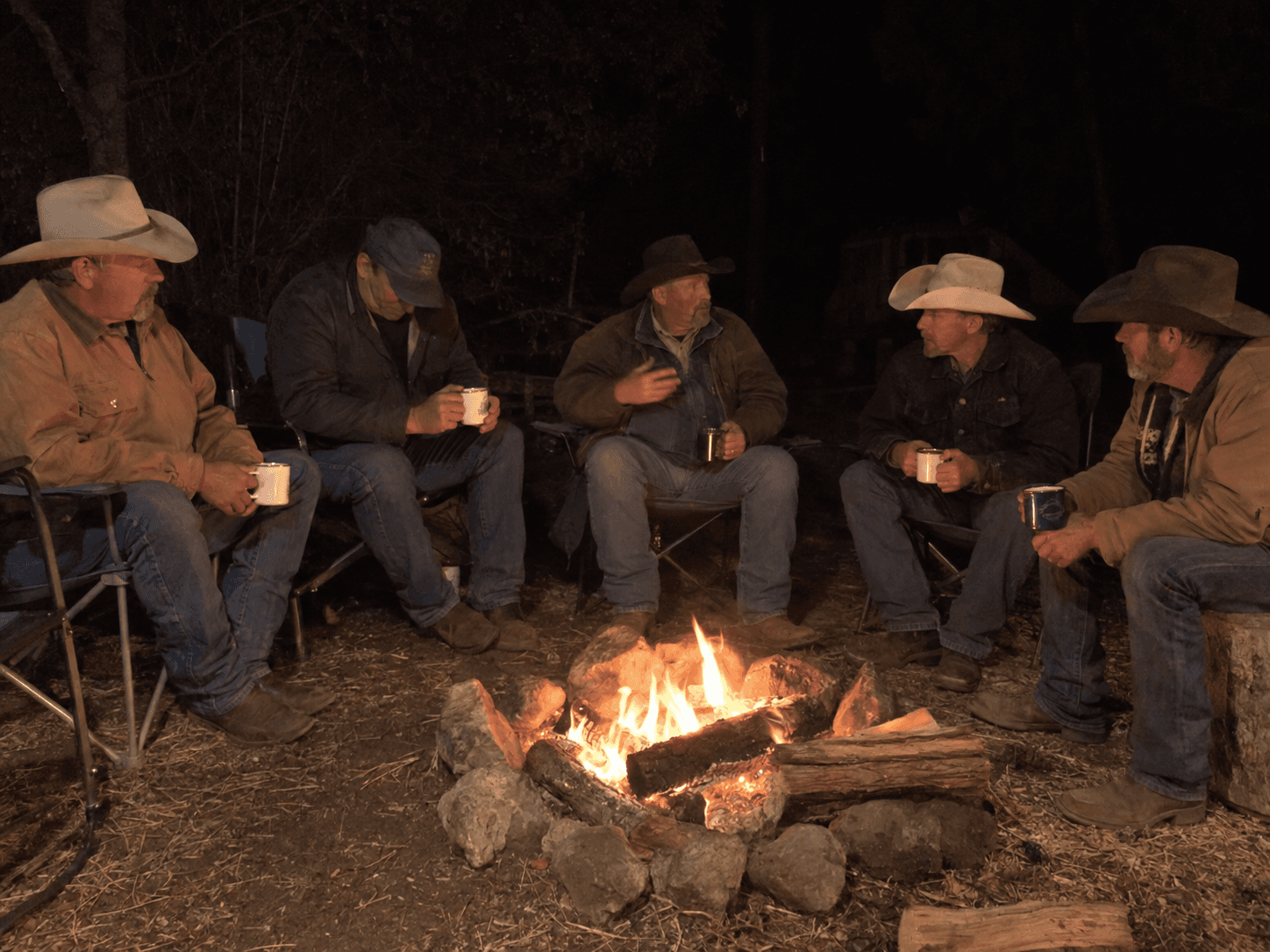 Five cowboys talking and drinking coffee around a campfire at night
